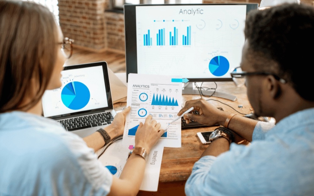 Dois profissionais analisando gráficos e relatórios de custos em uma mesa de trabalho