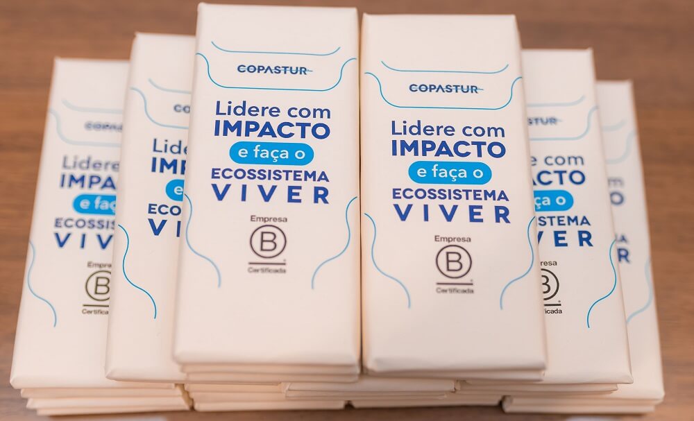 #pracegover: Chocolates embalados em papel reciclado bege, com detalhes azuis, logotipo da Copastur no topo e do Sistema B Brasil no rodapé, e frase no meio que diz "Lidere com impacto e faça o ecossistema viver"