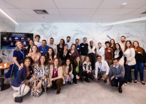 Café B na Copastur: futuro da liderança orientada ao impacto no Brasil (evento do Sistema B) - #pracegover: Pessoas participantes reunidas e sorridentes em frente à televisão com nomes e fotos de palestrantes do painel
