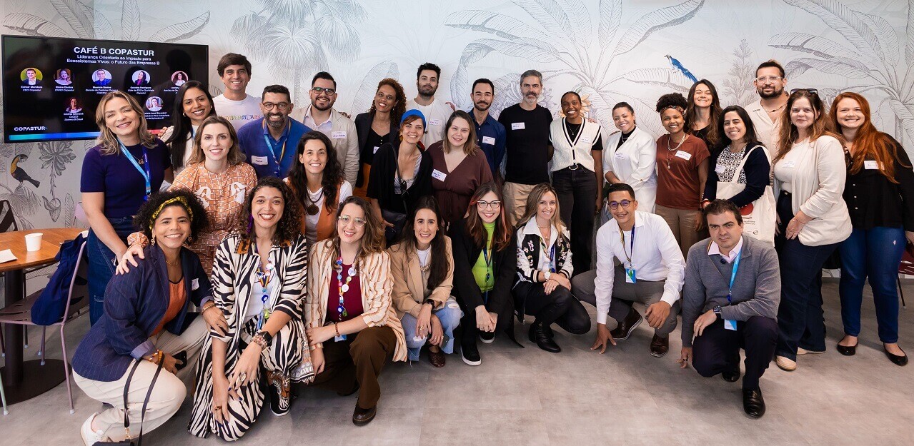Café B na Copastur: futuro da liderança orientada ao impacto no Brasil (evento do Sistema B) - #pracegover: Pessoas participantes reunidas e sorridentes em frente à televisão com nomes e fotos de palestrantes do painel