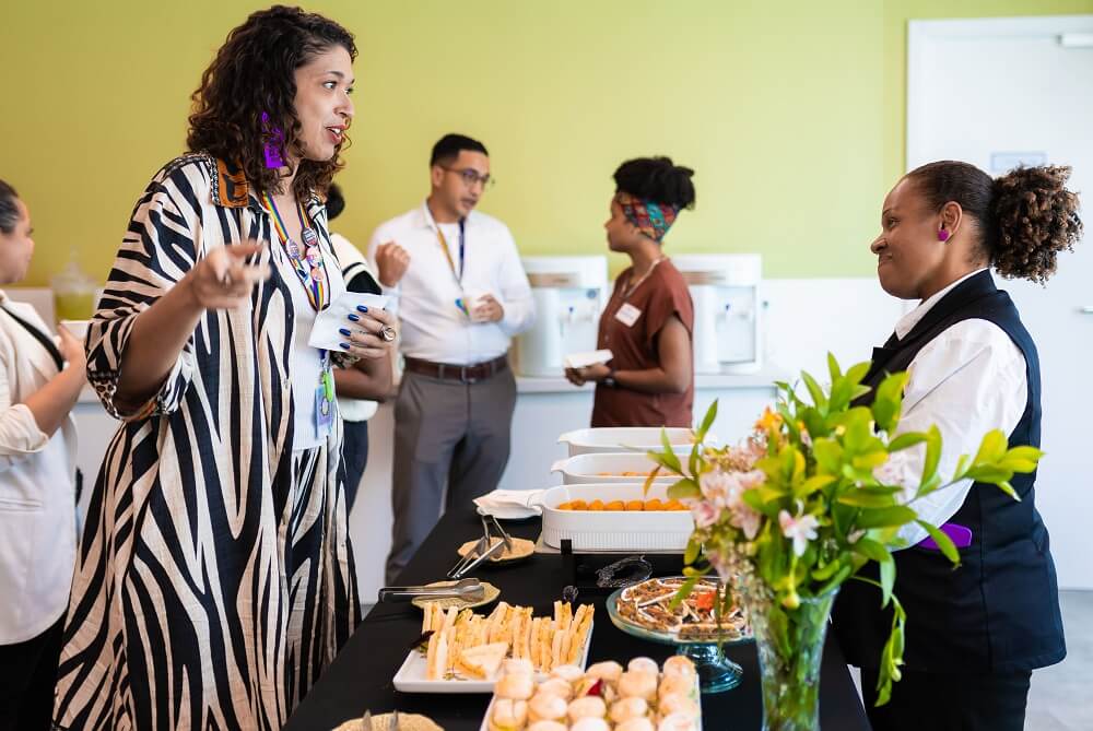 #pracegover: Pessoas conversam em volta da mesa de café da manhã e mulher mediadora do painel (Adrielly Honório), com cabelos escuros, curtos e cacheados, vestindo roupa com estampa de zebra, conversa com a garçonete do buffet, que tem cabelos escuros, cacheados e presos em um cabo de cavalo e veste uma blusa branca com colete preto