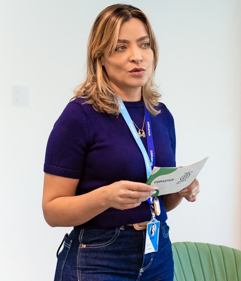 Elaine Dantas - CHRO Copastur - Café B (#pracegover: Mulher loira com cabelos na altura do ombro, vestindo camiseta azul escuro e crachá azul, segura ficha de palco com logotipo da Copastur e do Sistema B)