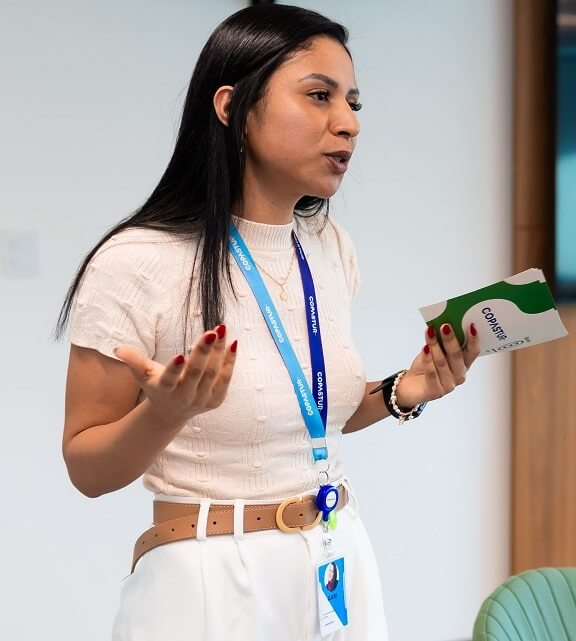 Gabriela Rodrigues - Líder de ESG e Qualidade na Copastur - Café B (#pracegover: Mulher com cabelos escuros e longos, vestindo camiseta bege com crachá azul, segura ficha de palco com logotipo da Copastur e do Sistema B)
