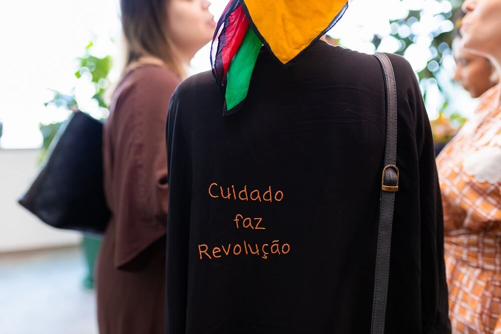 #pracegover: três mulheres conversam, e uma delas está de costas na foto vestindo uma blusa preta com escrito laranja que diz "cuidado é revolução", com alça da bolsa preta no ombro e bandana laranja, verde e vermelha