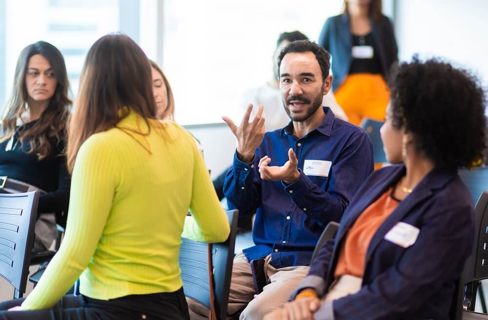 Participante do Café B faz pergunta para painelistas (#pracegover: homem de cabelos curtos e escuros, sentado na cadeira e usando camisa social azul escura, com adesivo colado escrito Well, fala e gesticula; em volta, outros participantes observam)