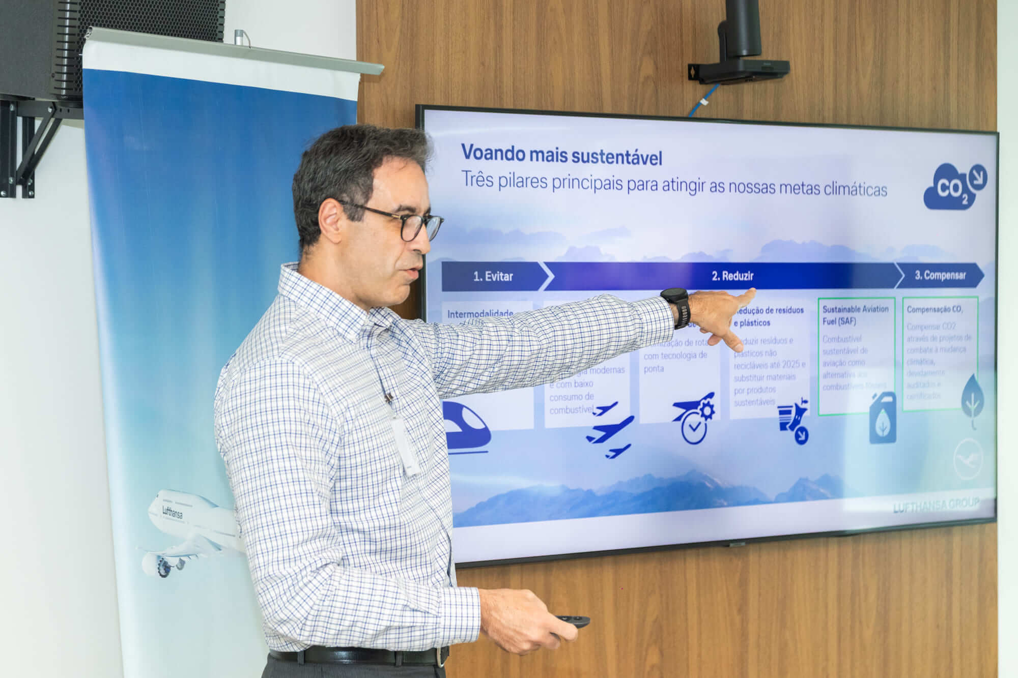 #PraCegoVer: Alan Jacch, homem de pele branca, óculos, cabelo curto escuro levemente grisalho, camisa xadrez branca com azul, calça social, cinto preto e relógio preto, está em pé diante de uma TV que mostra o título "Voando mais sustentável - Três pilares principais para atingir as nossas metas climáticas". Ele aponta com o braço estendido para a seção "2. Reduzir", que aparece em destaque azul no slide, entre 1. Evitar e 3. Compensar. O conteúdo inclui ações de sustentabilidade da companhia e ícones de trem, avião, check azul e símbolos relacionados a combustíveis sustentáveis, redução de resíduos e compensação de carbono. À esquerda, há um banner vertical azul com o desenho de um avião da Lufthansa.
