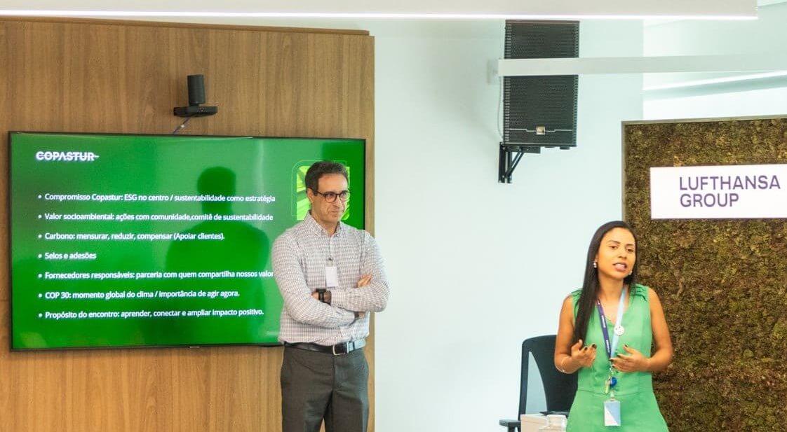 #PraCegoVer: Em uma sala de apresentação, Allan Jacch, homem de pele branca, cabelo curto e escuro levemente grisalho, óculos e camisa xadrez branca e azul está em pé, com os braços cruzados, ao lado de uma TV com slide verde com o logotipo da Copastur e, abaixo, os seguintes tópicos escritos em branco: “Compromisso Copastur: ESG no centro/sustentabilidade como estratégia. Valor socioambiental: ações com comunidade, comitê de sustentabilidade. Carbono: mensurar, reduzir, compensar (apoiar clientes). Selos e adesões. Fornecedores responsáveis: parceria com quem compartilha nossos valores. COP 30: momento global do clima/Importância de agir agora. Propósito do encontro: aprender, conectar e ampliar impacto positivo." À direita, Gabriela Rodrigues, mulher de pele negra, cabelos longos e lisos escuros e brincos prateados, veste um macacão verde com crachá da Copastur e fala para o público, gesticulando com as mãos. Ao fundo, uma parede verde revestida de plantas exibe uma placa iluminada com o logotipo da Lufthansa Group. Acima, há caixas de som instaladas na parede.