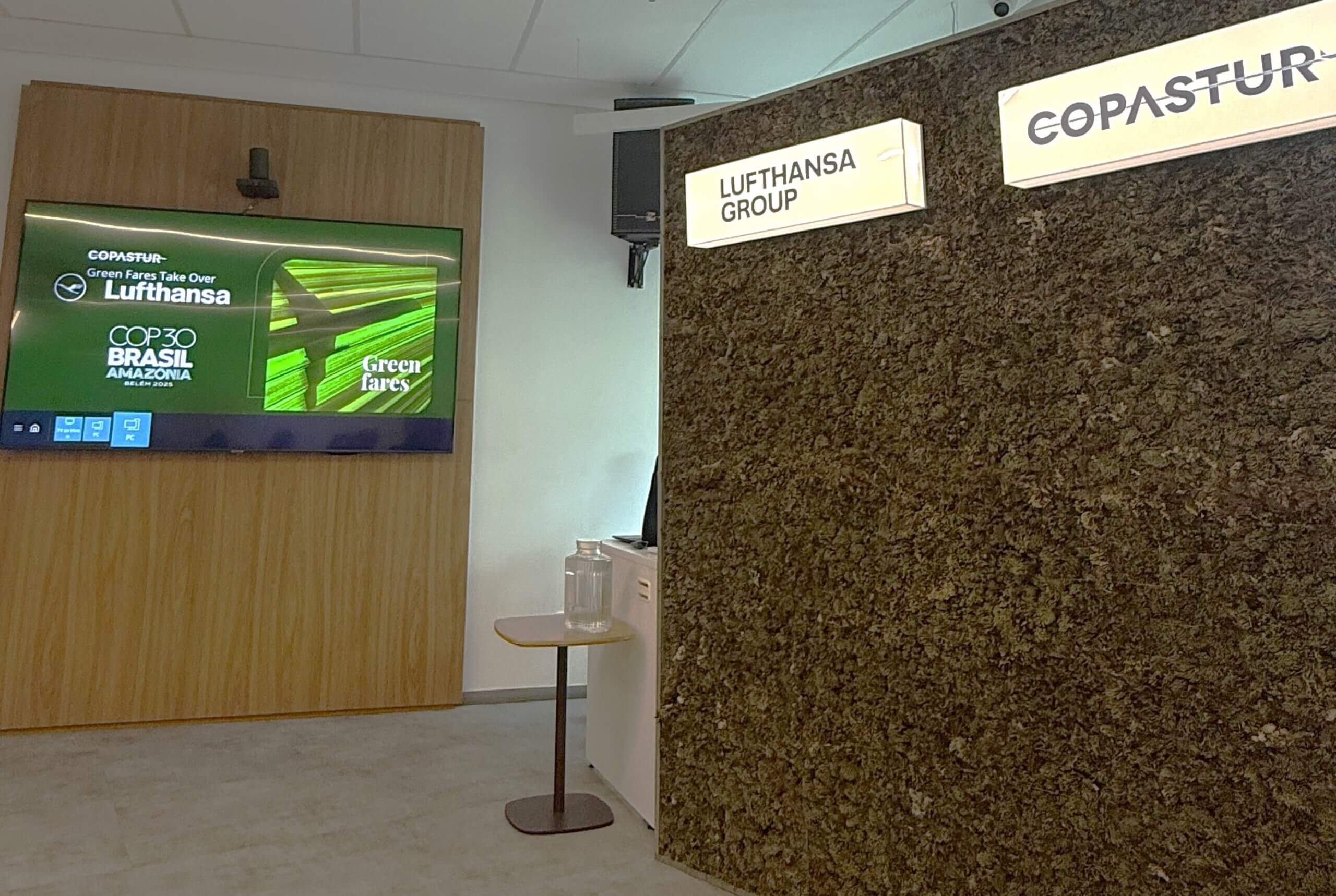 #PraCegoVer: Ambiente de apresentação corporativa, sala Brasilidades na Copastur. À esquerda, apoiada em uma parede de madeira, uma TV exibe um slide em tons de verde com os logotipos da Copastur e da Lufthansa e o texto "Green Fares Take Over", uma das ações de sustentabilidade em parceria, acompanhado do logotipo "COP 30 Brasil Amazônia Belém 2025" e de uma imagem aérea de plantações alinhadas com a sombra de um avião. No lado direito, há uma parede verde revestida de plantas, com duas placas iluminadas com os logotipos da Lufthansa Group e da Copastur, respectivamente. Em frente a essa parede, há uma mesa pequena com tampo claro e pés metálicos escuros, em que está apoiado um jarro transparente de água.