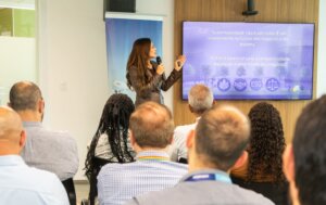 #PraCegoVer: Rosana Jatobá, mulher de pele branca, cabelos longos e lisos castanhos e blazer marrom fala para o público e olha para a tela, segurando um microfone. Ela está em frente a um banner com o céu azul e um avião da Lufthansa e diante de uma TV que exibe ícones de uma folha, um aperto de mãos, a sigla ESG e uma balança e, acima, as frases "Sustentabilidade não é um custo. É um investimento no futuro dos negócios e do planeta. O ESG é essencial para a competitividade, reputação e perenidade dos negócios”. A plateia aparece de costas, sentada em cadeiras pretas.