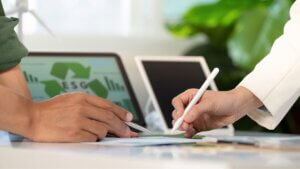 #PraCegoVer: duas mãos de mulheres, uma negra e uma branca, seguram canetas prateada e branca em cima de um papel; ao fundo, há um tablet com a tela verde e a sigla ESG e outro tablet desligado. (Práticas sustentáveis nas empresas: guia completo e case da Biosfera Copastur)
