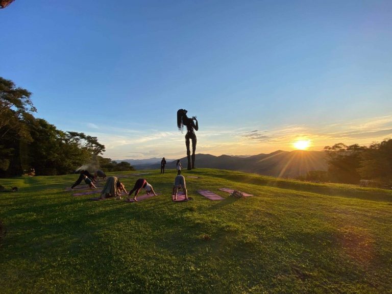 #PraCegoVer: Imagem ao ar livre no Ibiti Projeto, na Mata Atlântica mineira. Em um amplo gramado verde, no alto de uma colina, um grupo de pessoas pratica yoga sobre tapetes coloridos, todas inclinadas para a frente em posição de alongamento. Ao centro do espaço, há uma grande escultura de uma figura humana estilizada em pé. Ao fundo, a paisagem é composta por montanhas e áreas de floresta, com o sol se pondo no horizonte, iluminando o céu em tons de azul e dourado, criando uma atmosfera tranquila e contemplativa. (Descubra as viagens sustentáveis de incentivo da BioConexão - Foto: Divulgação/Ibiti Projeto)