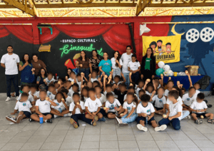 Fotografia de um grupo de crianças e pessoas adultas reunidas para uma foto coletiva no espaço cultural Cine Quebrada. As crianças estão sentadas no chão, em fileiras, usando principalmente camisetas brancas e calças escuras. Seus rostos estão desfocados para preservar a privacidade. Ao fundo, pessoas adultas estão em pé, sorrindo e posando para a foto. O espaço tem paredes decoradas com um mural artístico em vermelho, preto e azul, com referências ao cinema, incluindo ilustrações de rolos de filme. Na parede, aparece a inscrição “ESPAÇO CULTURAL”, acompanhada de um nome em letras verdes estilizadas. À direita, há uma faixa amarela com a ilustração de crianças e o nome “CINE QUEBRADA”, além de uma mesa azul com balões brancos e verdes. Acima, a estrutura do telhado tem vigas metálicas amarelas e telhas. O chão é revestido de grandes placas quadradas cinzas.