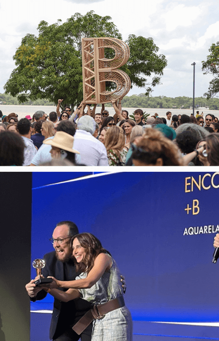Colagem vertical com duas fotografias relacionadas ao Encontro+B™ Amazônia 2025 e à premiação da Aquarela Agência. Na imagem superior, participantes do Encontro+B™ Amazônia 2025 se reúnem em uma área aberta cercada por árvores, próxima a um rio. No centro da foto, várias pessoas erguem uma grande letra “B” decorativa, confeccionada em material trançado de tom amadeirado. A cena é marcada pela presença de uma ampla multidão, vegetação abundante ao fundo e céu com muitas nuvens. Na imagem inferior, Raphael Sacchetto, Head da Aquarela Agência, e Marina Weisshaupt, Supervisora da Aquarela Agência, aparecem sobre um palco durante uma cerimônia de premiação. Ambos seguram o troféu do Prêmio Live 2026 enquanto posam sorrindo para a fotografia. Ao fundo, um painel azul exibe os dizeres “ENCONTRO +B” e “AQUARELA”. À direita da imagem, aparece parcialmente a mão de uma pessoa segurando um microfone. A cena registra o momento de reconhecimento da Aquarela Agência na premiação.