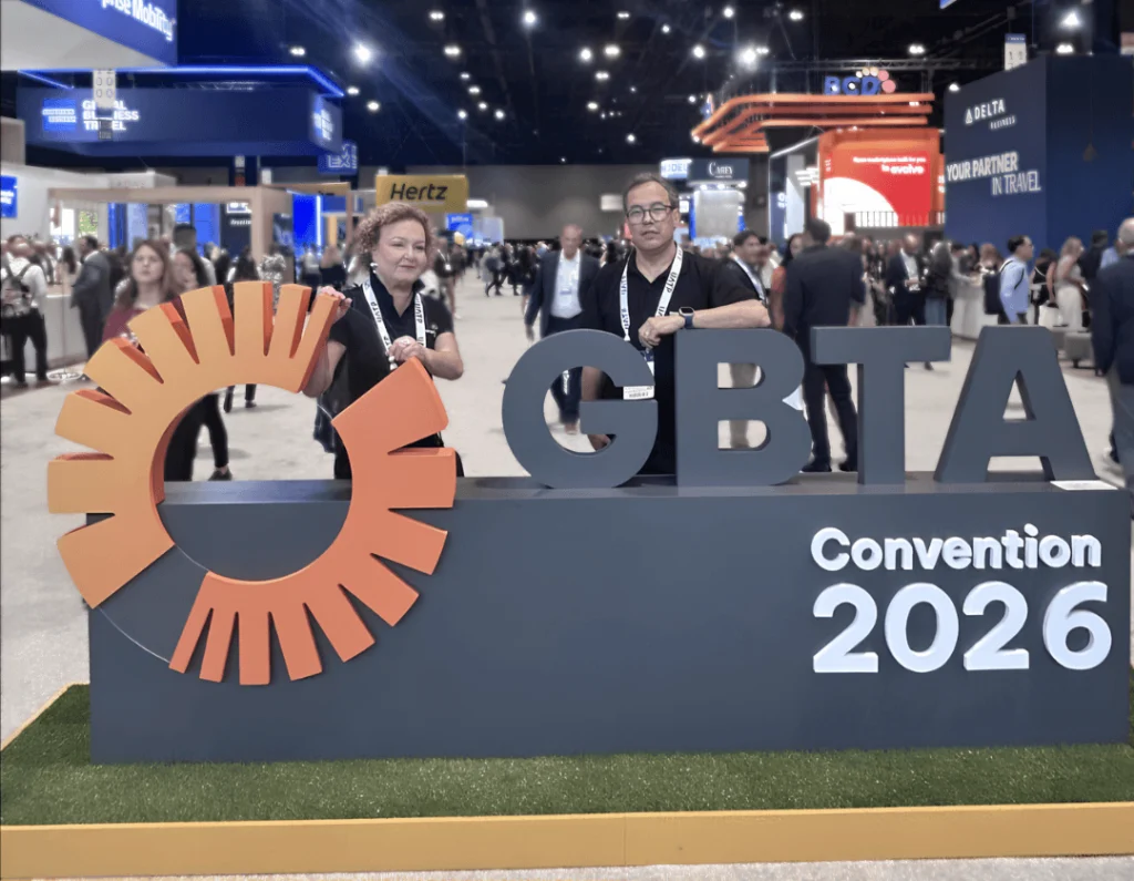 #PraCegoVer: Foto da GBTA Convention 2026, em um grande pavilhão de eventos. Em primeiro plano, há um palanque azul-escuro com o logotipo laranja da GBTA e os letreiros “GBTA” e “Convention 2026”. À esquerda, Lylian Poloni, uma mulher de pele clara, com cabelos curtos, cacheados e loiros, usa uma camisa polo escura, crachá preso a um cordão branco e brincos. Ela está apoiada no palanque e olha em direção à câmera. À direita, Felipe Mesquita, um homem de pele clara, com cabelos curtos e escuros, usa óculos de grau, camisa polo preta, crachá em um cordão branco e relógio. Ele também está encostado no palanque e olha para a câmera. Ao fundo, o pavilhão está movimentado, com diversas pessoas e estandes de empresas, parcialmente desfocados para manter o foco nas duas pessoas e no palanque.