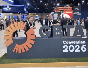 #PraCegoVer: Foto da GBTA Convention 2026, em um grande pavilhão de eventos. Em primeiro plano, há um palanque azul-escuro com o logotipo laranja da GBTA e os letreiros “GBTA” e “Convention 2026”. À esquerda, Lylian Poloni, uma mulher de pele clara, com cabelos curtos, cacheados e loiros, usa uma camisa polo escura, crachá preso a um cordão branco e brincos. Ela está apoiada no palanque e olha em direção à câmera. À direita, Felipe Mesquita, um homem de pele clara, com cabelos curtos e escuros, usa óculos de grau, camisa polo preta, crachá em um cordão branco e relógio. Ele também está encostado no palanque e olha para a câmera. Ao fundo, o pavilhão está movimentado, com diversas pessoas e estandes de empresas, parcialmente desfocados para manter o foco nas duas pessoas e no palanque.