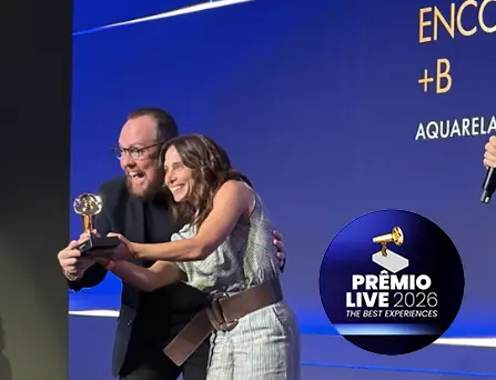 Fotografia de uma cerimônia de premiação em um palco com painel azul-escuro e iluminação azul e branca. Um homem branco de óculos, cabelos escuros e barba curta e uma mulher branca de cabelos castanhos comemoram no palco, sorrindo enquanto seguram um troféu dourado sobre uma base preta. O homem veste roupa preta, e a mulher usa vestido prateado de brilho metálico, com cinto largo marrom. Ao fundo, há um painel azul-escuro com iluminação azul e branca. No canto superior direito, aparece um selo circular azul-marinho com o texto “PRÊMIO LIVE 2026 – THE BEST EXPERIENCES”.